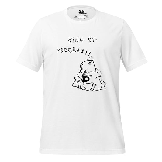 The image is a white t-shirt with a drawing of a capybara wearing a blanket and holding a mug, looking cosy. The design features the words "KING OF PROCRASTINATION". Unruly Folk brand.