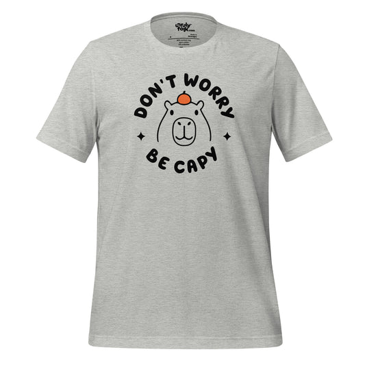 The image is of an athletic heather t-shirt with a slogan that says "Don't Worry Be Capy". It has a line drawing of a capybara with an orange on its head. From the Unruly Folk brand.